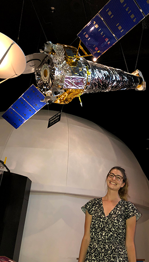 Adi Foord standing beneath a model of the Chandra X-ray Observatory