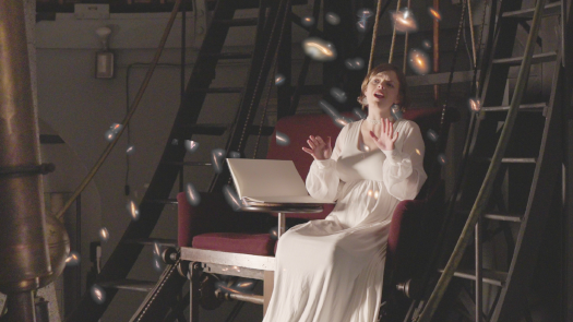 A young woman in a white dress is seated in a chair in an observatory. She is singing and emoting with her hands. Images of semi-transparent galaxies are superimposed on the scene.